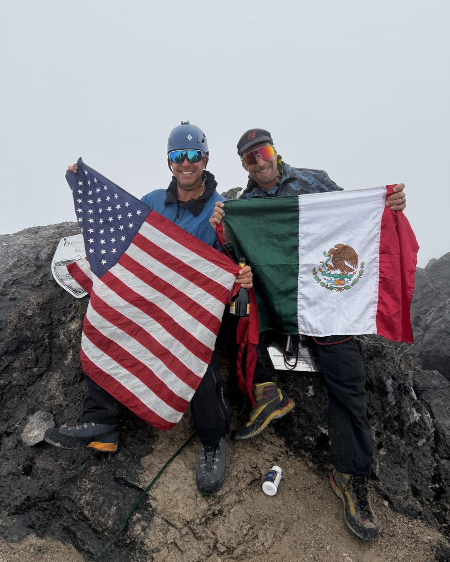 Mark Parella and a fellow climber on the summit of Carstensz Pyramid in 2026, holding the flags of the United States and Mexico