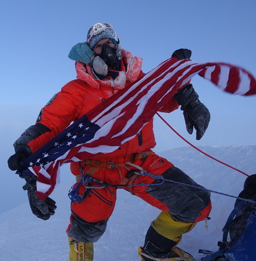 Mark Parella holding an American flag on the summit of Mount Everest in 2019, wearing an oxygen mask