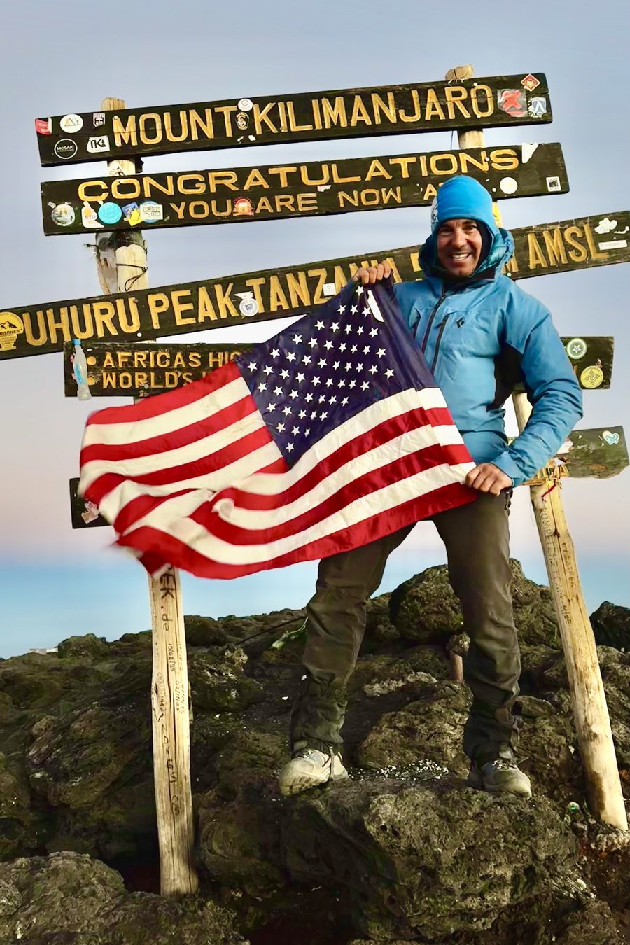Mark Parella holding an American flag at the Uhuru Peak summit sign on Mount Kilimanjaro in 2026