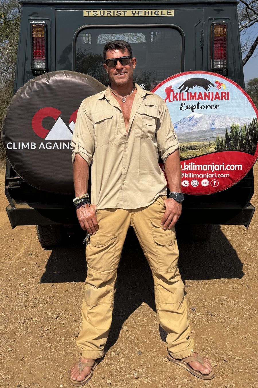 Mark Parella in safari clothes standing in front of a Kilimanjari Explorer safari vehicle with a Climb Against Cancer tire cover, Serengeti 2026